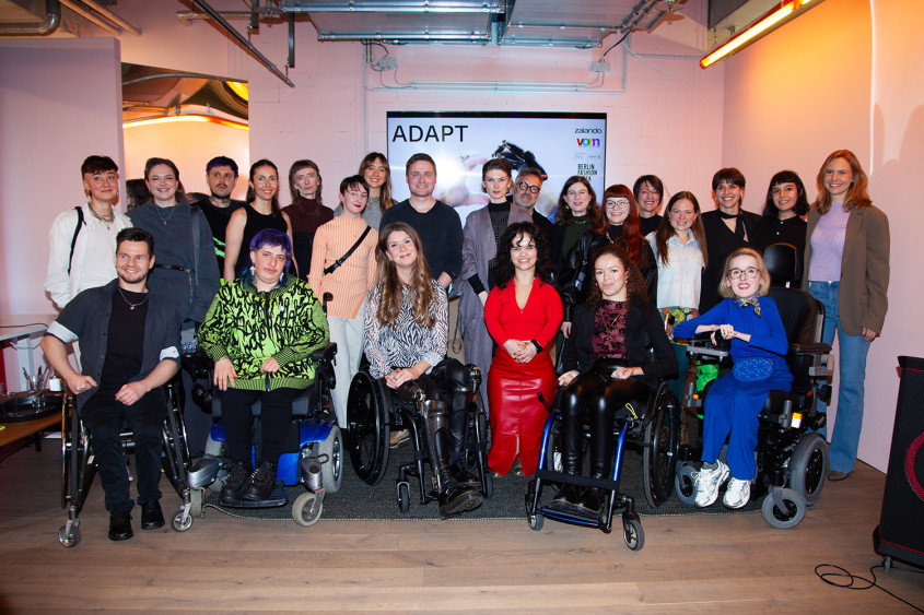 A group of about twelve people poses together smiling in front of a white wall banner reading: vorn x zalando Design academy ADAPT. The group features diverse individuals, including people using wheelchairs and a woman with dwarfism wearing a bright red outfit in the center foreground.