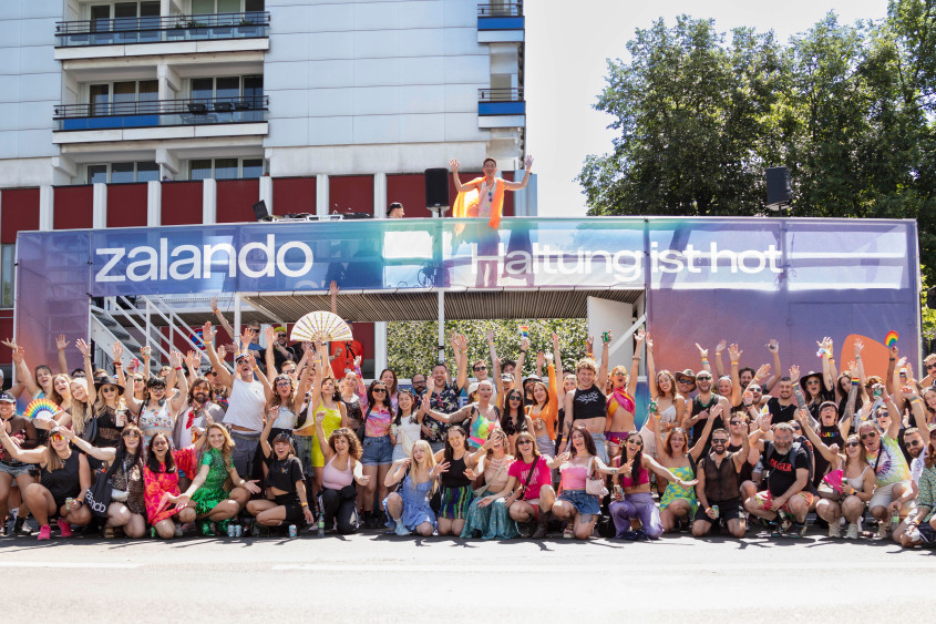 Wide shot of a massive, diverse crowd gathered on a street, cheering and waving. In the background is a large purple and blue float with white text that reads "zalando" and "Haltung ist hot". People are celebrating, some holding a fan. One person is waving from the roof of the float.