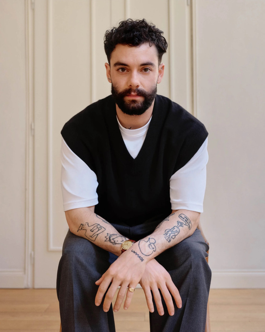 A bearded man leaning forward with his arms crossed, wearing a white tshirt and a black debardeur