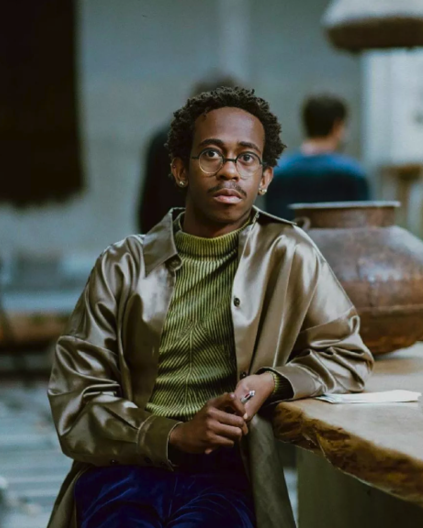 a Black man with glasses, wearing a green striped top and a silk shirt on top