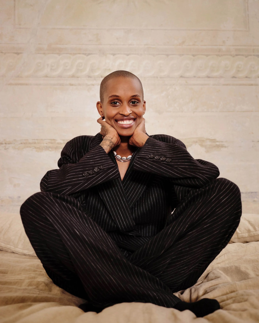 a Black woman with a shaven head, wearing a black oversized outfit is sitting cross-legged on a bed