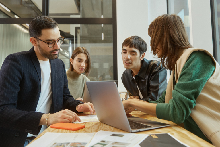 A group of diverse people sitting around a table discussing something on a laptop