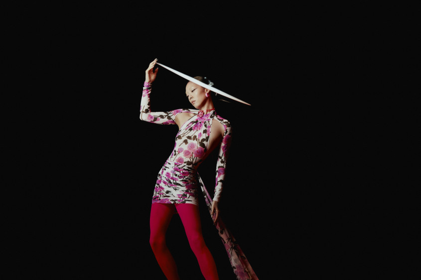 Rina Sawayama standing with a pink dress, red tights and a large hat for Zalando